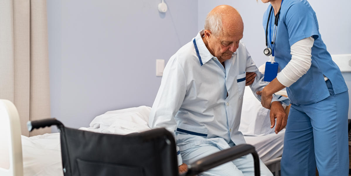 Lovely nurse assisting senior man to get up from bed at hospital. Caring nurse supporting elderly patient while getting up from bed and move towards wheelchair at nursing home. Disabled man standing up at care centre while caregiver helping him to get up in the morning.